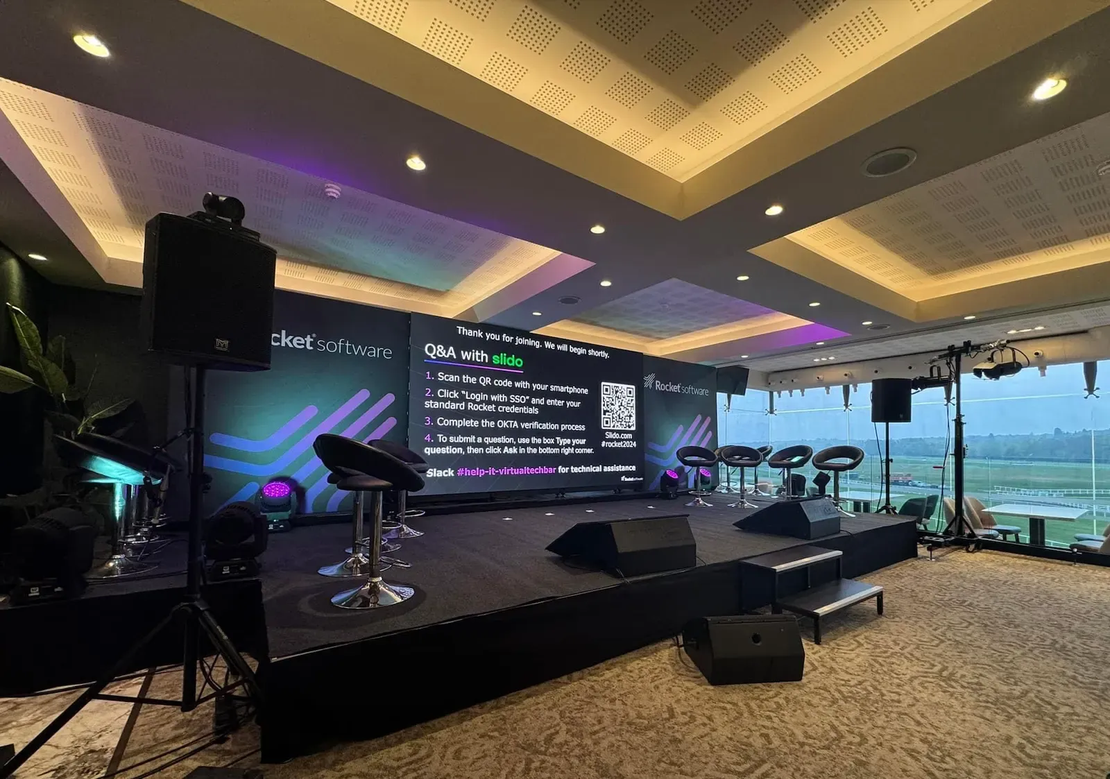 A corporate conference stage with an LED wall showing a Q&A slide, presenter stools, moving lights and a camera on a stand